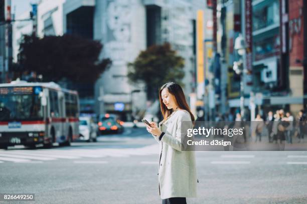 young woman using smartphone on city street while waiting for taxi, against busy transportation and commuters - distrito de shibuya fotografías e imágenes de stock