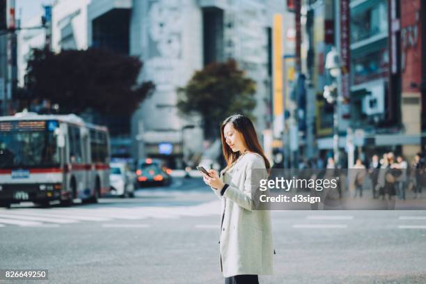 young woman using smartphone on city street while waiting for taxi, against busy transportation and commuters - shibuya station foto e immagini stock