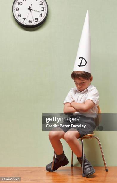 humiliated schoolboy sitting in classroom with dunces hat on - school punishment stockfoto's en -beelden