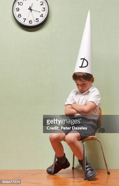 humiliated schoolboy sitting in classroom with dunces hat on - dumstrut bildbanksfoton och bilder