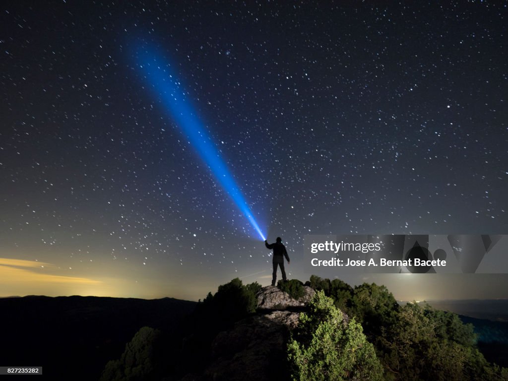 Silhouette of a man on the top of a mountain in the night, with a lantern in the hand doing a beam of light on a sky of stars