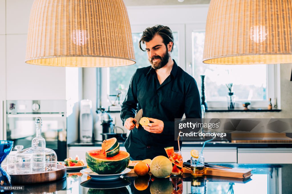 Young bearded man enjoying watermelon and making salad in the kitchen