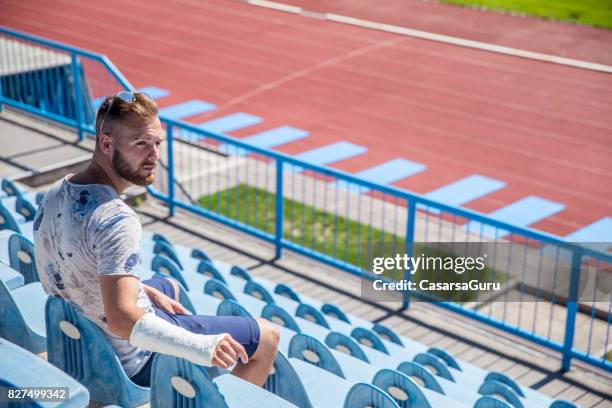 junger erwachsener mann mit gebrochenen arm auf der tribüne sitzen - armbruch stock-fotos und bilder
