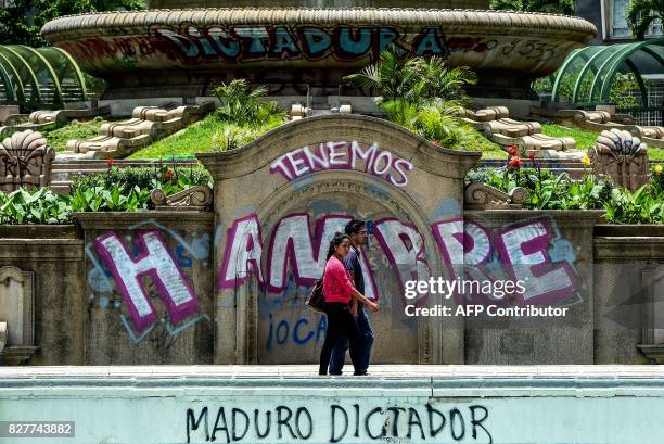 Couple walks by graffitis reading "We Are Hungry" and "Maduro Dictator" in Caracas on August 8, 2017. Recent demonstrations in Venezuela have stemmed...