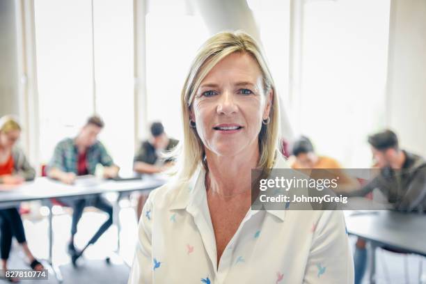 mature female teacher looking at camera, college students working in background - teacher portrait stock pictures, royalty-free photos & images