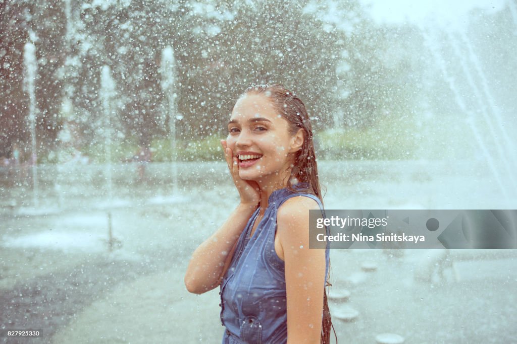 Laughing young woman in wet clothes standing in water splashes of fountain