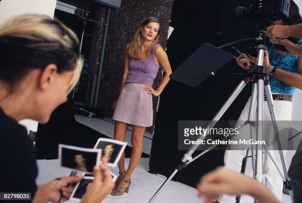 British Supermodel Kate Moss, center, stands with hand on hip as an unidentified makeup artist, foreground, looks through Polaroids during a fashion...