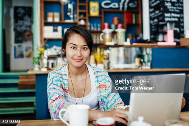 young woman smiling while working on laptop in colourful coffee shop - indonesian ethnicity foto e immagini stock