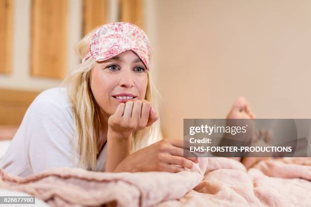 beauty woman with long blonde hair lying down in bed in stylish hotel room with wooden planks hanging from the wall, she is wearing a bathrobe and a sleep mask on forehead while watching tv enjoying me time, man sleeps beside her, we see his feet only. - woman lying on stomach with feet up fotografías e imágenes de stock