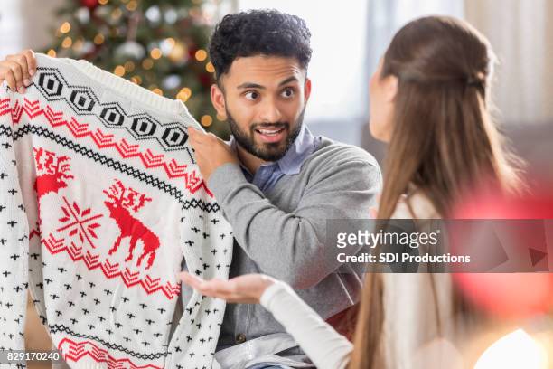 man with funny expression holds up christmas sweater - ugliness stock pictures, royalty-free photos & images