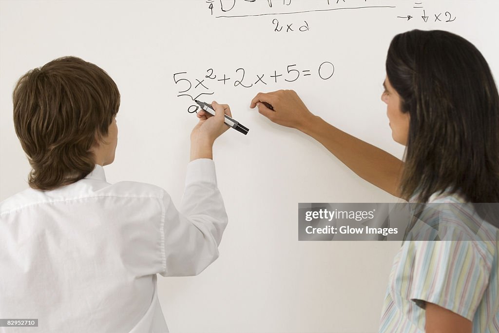 Rear view of a teenage boy solving algebra on whiteboard and a female teacher standing beside him