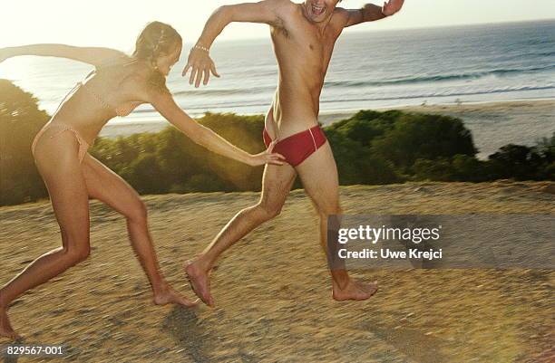 woman chasing man, pulling at swimming trunks - young men in speedos 個照片及圖片檔