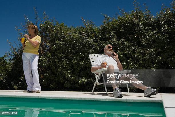 senior man talking on a mobile phone and senior woman drinking juice at poolside - klappstuhl stock-fotos und bilder