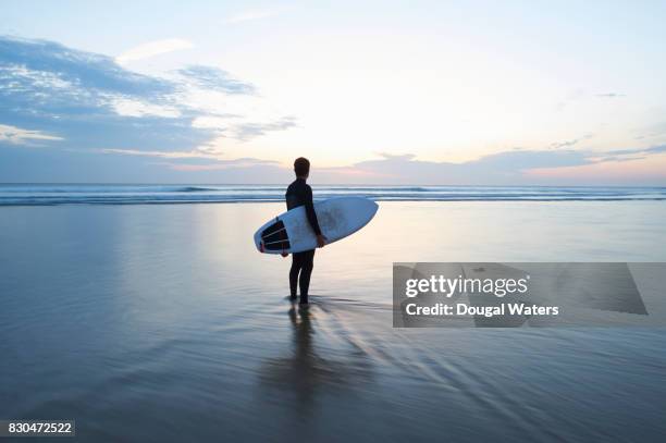 surfer with surfboard looking out to sea at dusk. - beach hold surfboard stock-fotos und bilder