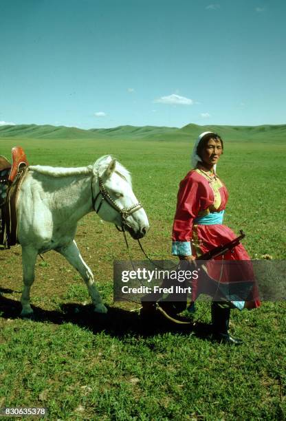 Mongolian nomad woman walking with her horse after practicing her rifle skills in the grassland. She belongs to the nomads police. The deel outfit is...