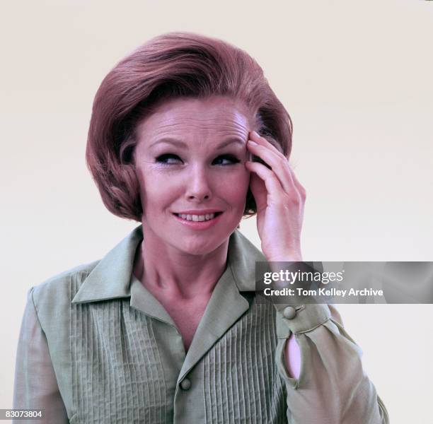 Portrait of a red-headed woman in a green blouse as she puts her hand to her forehead as though she has a headache and smiles unconvincingly,...