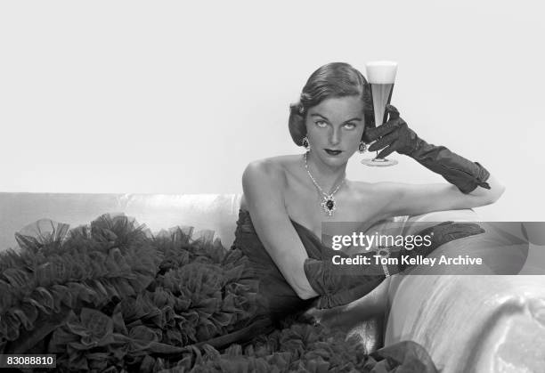 Fashionably dressed in a strapless, taffeta gown, elbow gloves, and wearing a jeweled necklace and earrings, a women reclines on a sofa and holds a...