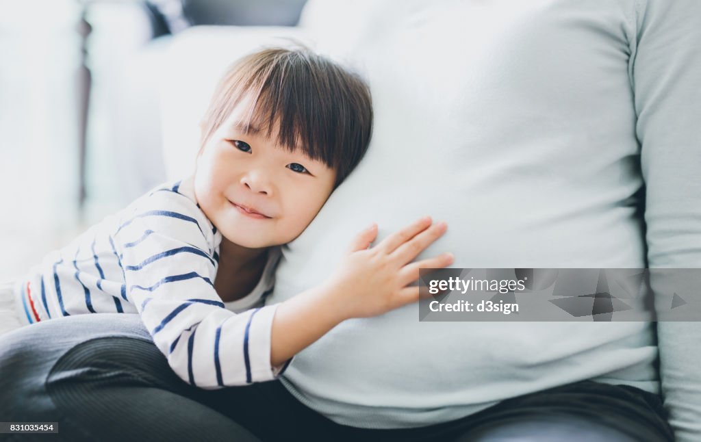 Cute little Asian girl touching and listening to pregnant mother's belly with joy