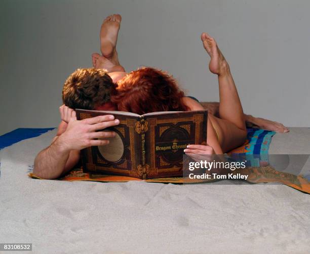 An unidentified man and a woman in swimwear lie together on a blanket on a sandy beach set in a studio, mostly hidden behind a copy of an oversize...