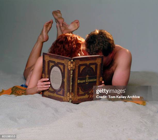 An unidentified man and a woman in swimwear lie together on a blanket on a sandy beach set in a studio, mostly hidden behind a copy of an oversize...