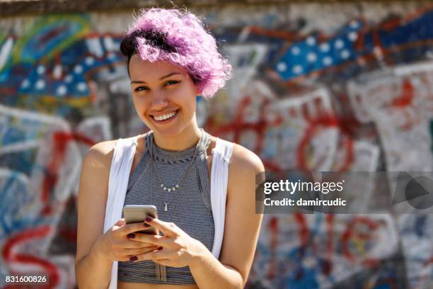 smiling young hipster girl using smart phone against graffiti wall - colourful graffiti stock pictures, royalty-free photos & images