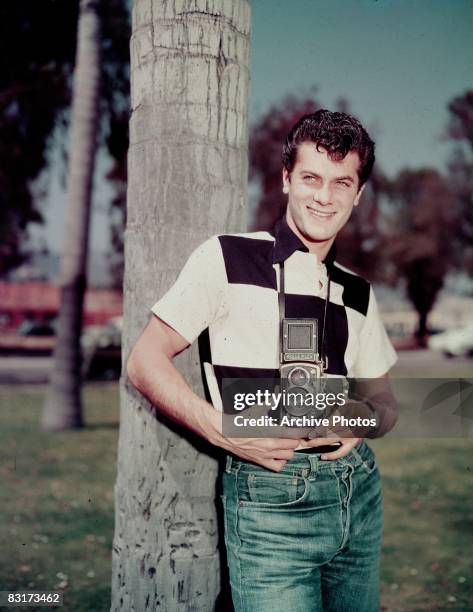 American actor Tony Curtis leans against a tree, holding a Rolleiflex camera, 1950s.