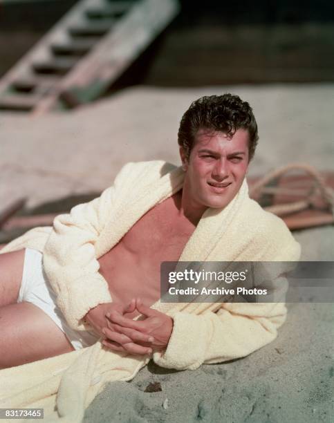 American actor Tony Curtis reclines on the beach in a terry robe and white swimming trunks, 1950s.