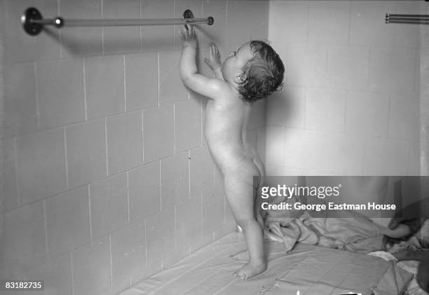 Full length portrait of a baby boy reaching up to the towel bar while being bathed, United States, ca.1920s.