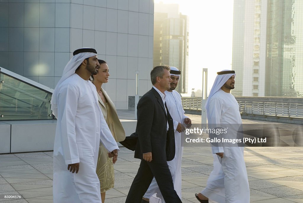 Businessman and woman walking with men dressed in traditional Middle Eastern attire, Dubai cityscape in background, UAE