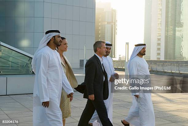 businessman and woman walking with men dressed in traditional middle eastern attire, dubai cityscape in background, uae - dubai difc stock pictures, royalty-free photos & images