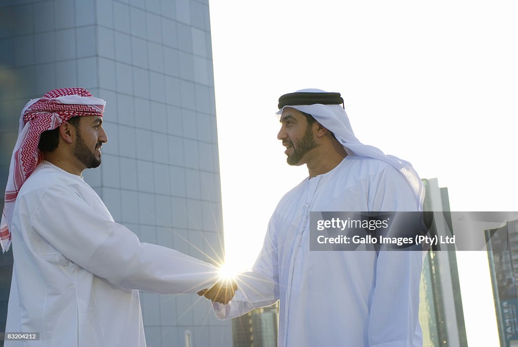 Men dressed in traditionally Middle Eastern attire shaking hands, Dubai cityscape in background, UAE