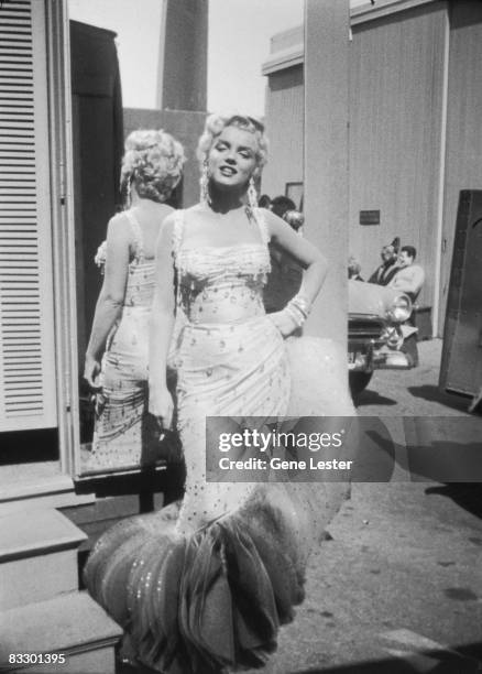 Portrait of American actor Marilyn Monroe , in a light-colored, beaded gown, as she stands with her back to a mirror, a cigarette in one hand and the...