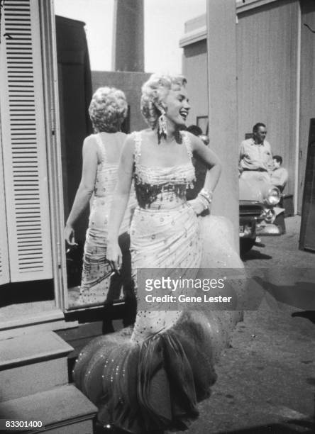 Portrait of American actor Marilyn Monroe , in a light-colored, beaded gown, as she laughs with her back to a mirror, a cigarette in one hand and the...