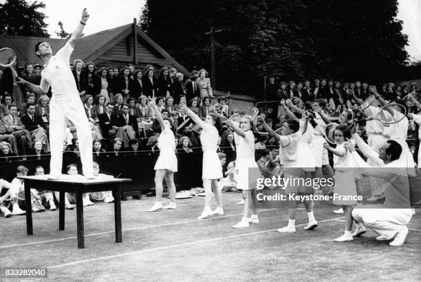 Dan Maskell debout sur la table expliquant les gestes et mouvements avec une raquette de tennis pendant que Fred Perry aide les élèves, le 6 juillet...