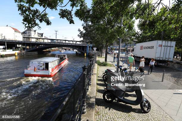 Sightseeing barge sails past a row of Coup eScooter electric hire vehicles, operated by Robert Bosch GmbH, as they stand on a bank of the River Spree...