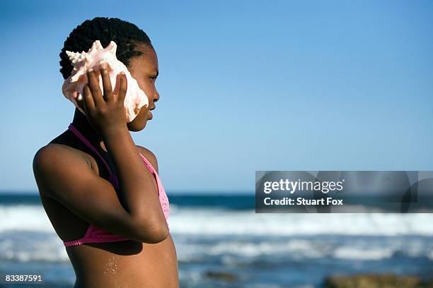 young girl holds giant shell against her ear as she looks out at the indian ocean, kwazulu natal province, south africa - zulu girls - fotografias e filmes do acervo