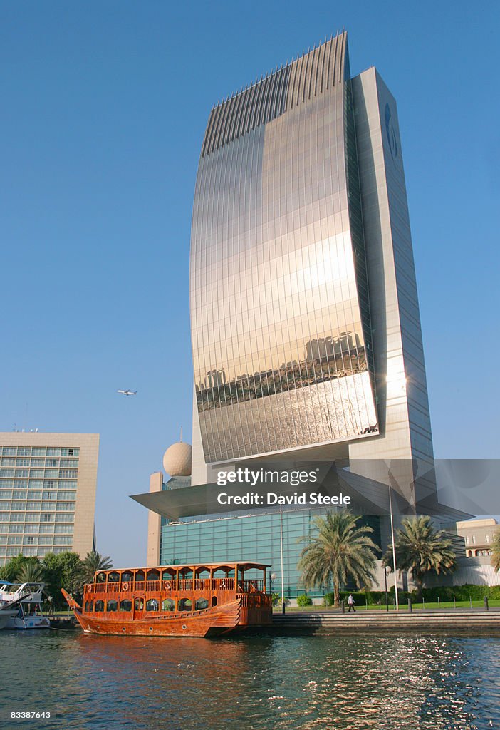 The National Bank of Dubai building with a wooden restaurant dhow moored in front of it, City of Dubai, Emirate of Dubai, United Arab Emirates.