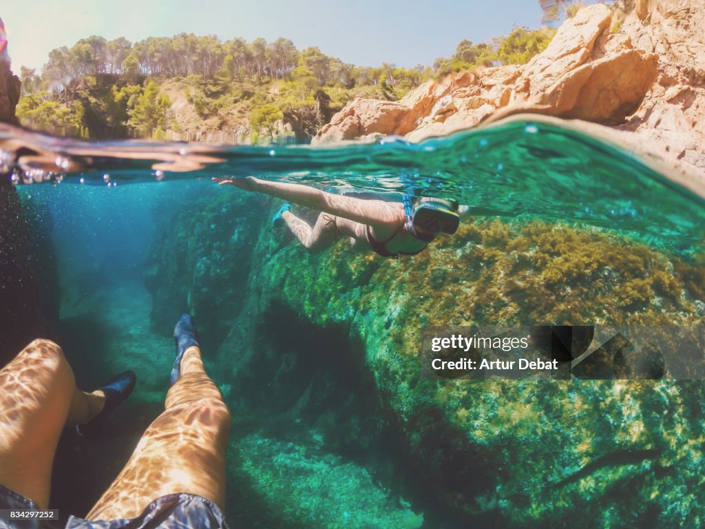 Couple doing snorkel exploring the natural cave in the shoreline of Costa Brava Mediterranean Sea during summer vacations in a paradise place taking picture with dome cover and underwater view.