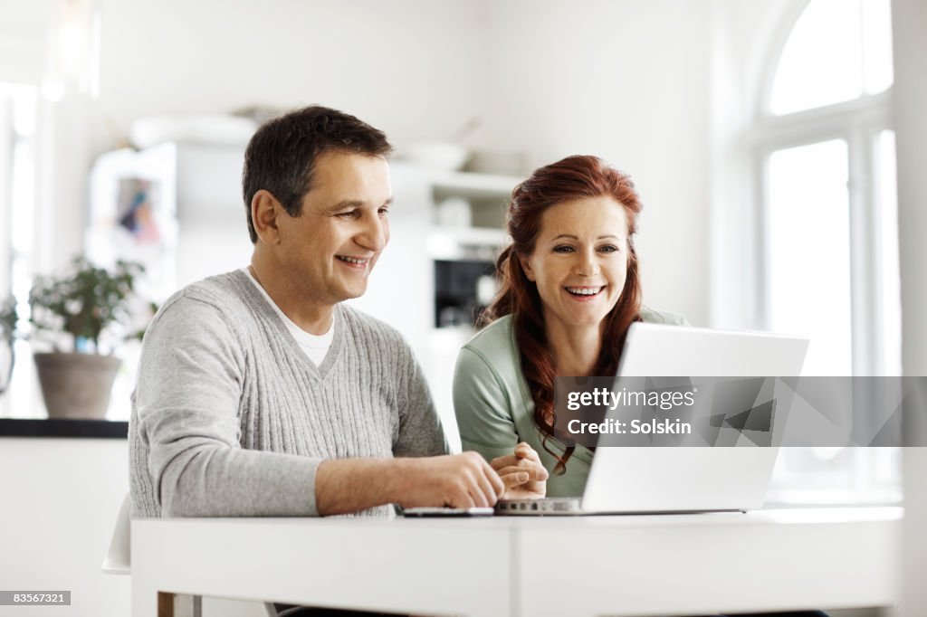 Mature couple sitting in kitchen, using laptop