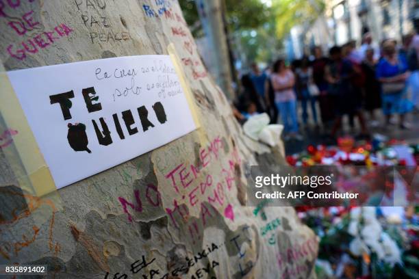 Message reading "I love you" is displayed on a tree a people stand next to flowers, candles and other items set up on the Las Ramblas boulevard in...