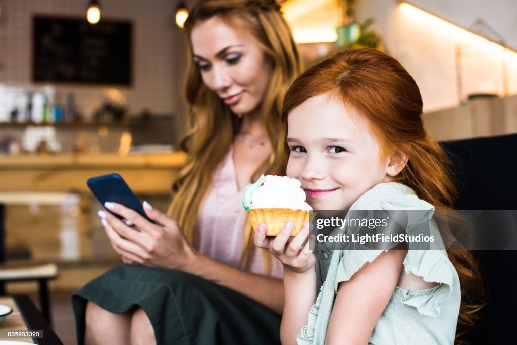Linda chica pelirroja sonriente comer Magdalena mientras que madre con smartphone detrás