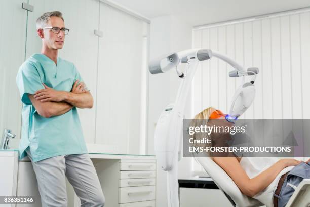 aesthetic cosmetic dentistry dental care - beautiful woman lying on dentist’s office chair with mouth open for tooth bleaching while wearing protective glasses and lip retractor is inserted the whitening unit is emitting ultraviolet light. - retractor stock pictures, royalty-free photos & images