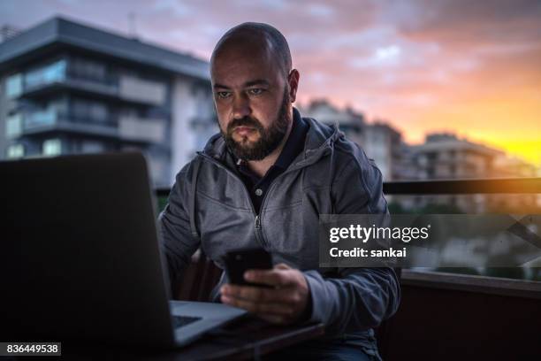 禿頭和大鬍子的男人在日落時使用筆記本電腦和智慧手機 - suspicion 個照片及圖片檔