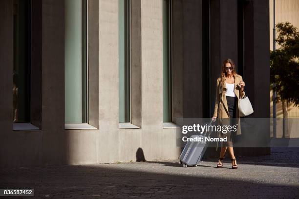 businesswoman with luggage walking outside airport - trench coat stock pictures, royalty-free photos & images