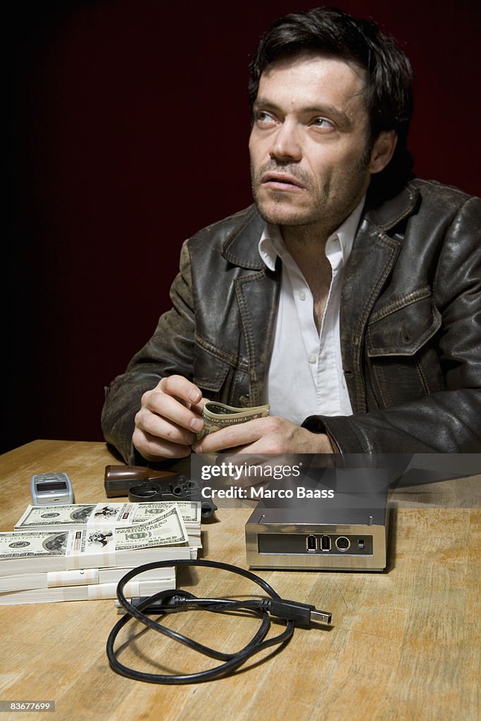 A man sitting at a table with stacks of money, a mobile phone and a hard drive