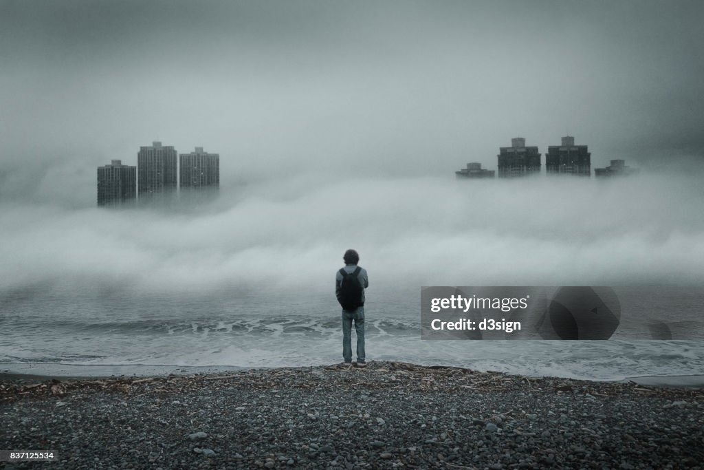 Man standing alone looking out to sea against moody sky during foggy weather