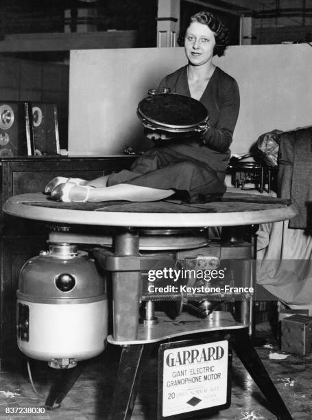 Jeune femme assise sur un énorme moteur de phonographe électrique tient dans ses mains un modèle standard de phonographe, à Londres, Royaume-Uni en...