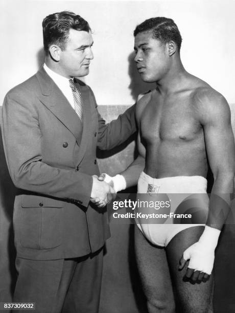 Le boxeur Jimmy Braddock en costume de ville serre la main au boxeur noir américain Joe Louis en sous-vêtement lors de l'entraînement de ce dernier...
