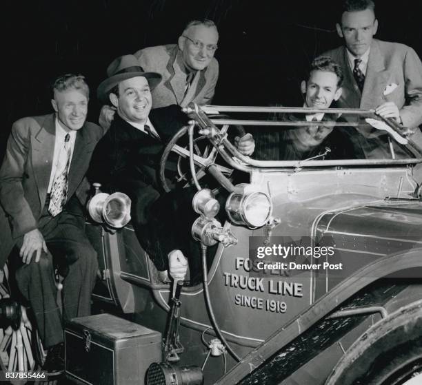 James Melton, famed tenor and radio star and avid antique automobile fan, grasps the handbrake of an old model as several members of the Horseless...