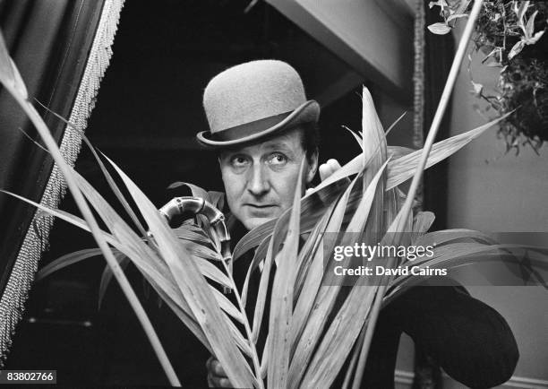 English actor Patrick Macnee concealed behind a houseplant, 20th May 1965.